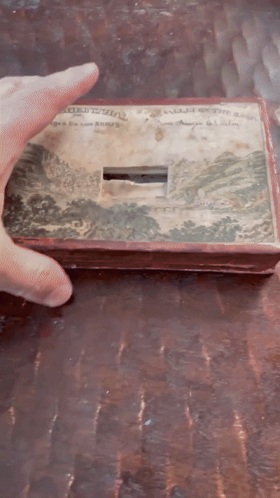 An animated GIF that shows someone opening up an old fashioned book that is very antique. When they open it up there is an accordion of paper between the front cover and back cover, And the animated GIF shows a little hole cut in the cover of the book. When we peer through that hole we see a diorama sneaking off at the distance that looks like mountains of a countryside far away. Than the animated GIF shifts to a close up of The interior of the book, and we can see that is a parallax effect, You can see one part of the countryside — a hill with a bridge — with towers and towns further in the distance, and as the camera view tilts slightly from side to side, the closer scenes appear to "move" more quickly than the ones in the back