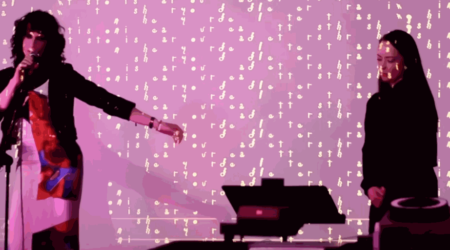 a poet and an interpreter perform in moody lighting. lattice-like patterns of letters are projected onto them and the wall behind.