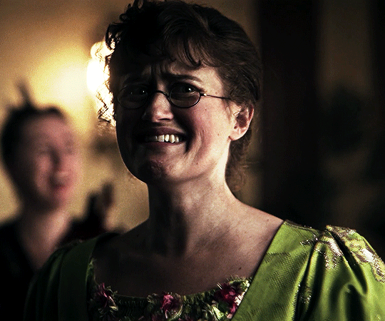 A gif of Mary Bennet from The Other Bennet Sister, in an olive green dress with her hair up, wearing her spectacles and smiling sheepishly while she looks around the room. Someone in the background is applauding her.