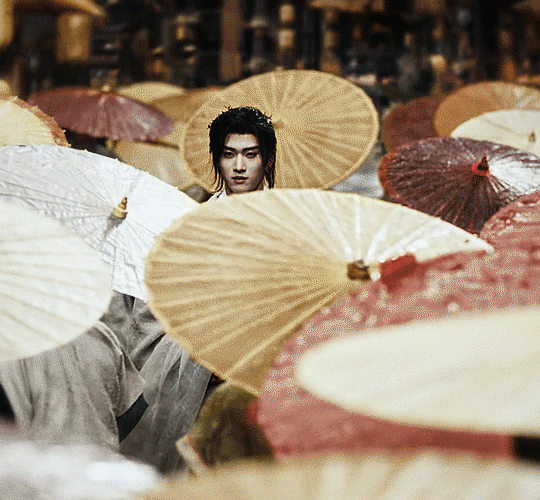 gif from the TV show Fangs of Fortune, featuring a young demon Li Lun in white robes, holding a magical rattle. It's raining on him while he's alone but surrounded by dozens of humans all with umbrellas
